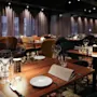 Borde dækket med vinglas, tallerkener og servietter, der skaber en elegant spisestemning i en svagt oplyst restaurant med mønstret tæppe, farverige stole og gardinbeklædte vinduer.