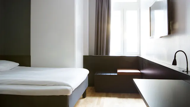 En säng är prydligt bäddad i ett minimalistiskt rum med en väggmonterad TV och ett soligt fönster inramat av mörka gardiner, vilket skapar en ljus och enkel atmosfär.