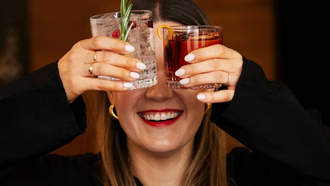 En person med langt hår holder to cocktails foran ansiktet, dekker det. Ett glass har en klar væske og rosmarin; det andre har en rød drikk. Bakgrunnen er uskarp.