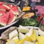 Vannmelon og blandede grønnsaker ligger i boller på et bord, omgitt av skiver av agurk, tomater og grønnsaker. Bakgrunnen har flere skivede frukter og kjøtt. Avispapir er synlig.
