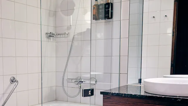 Et badekar med en dusjhode er omgitt av hvite flislagte vegger og en glassvegg. Ved siden av er en mørk benkeplate med en servant. Flere toalettartikler er montert på veggen.