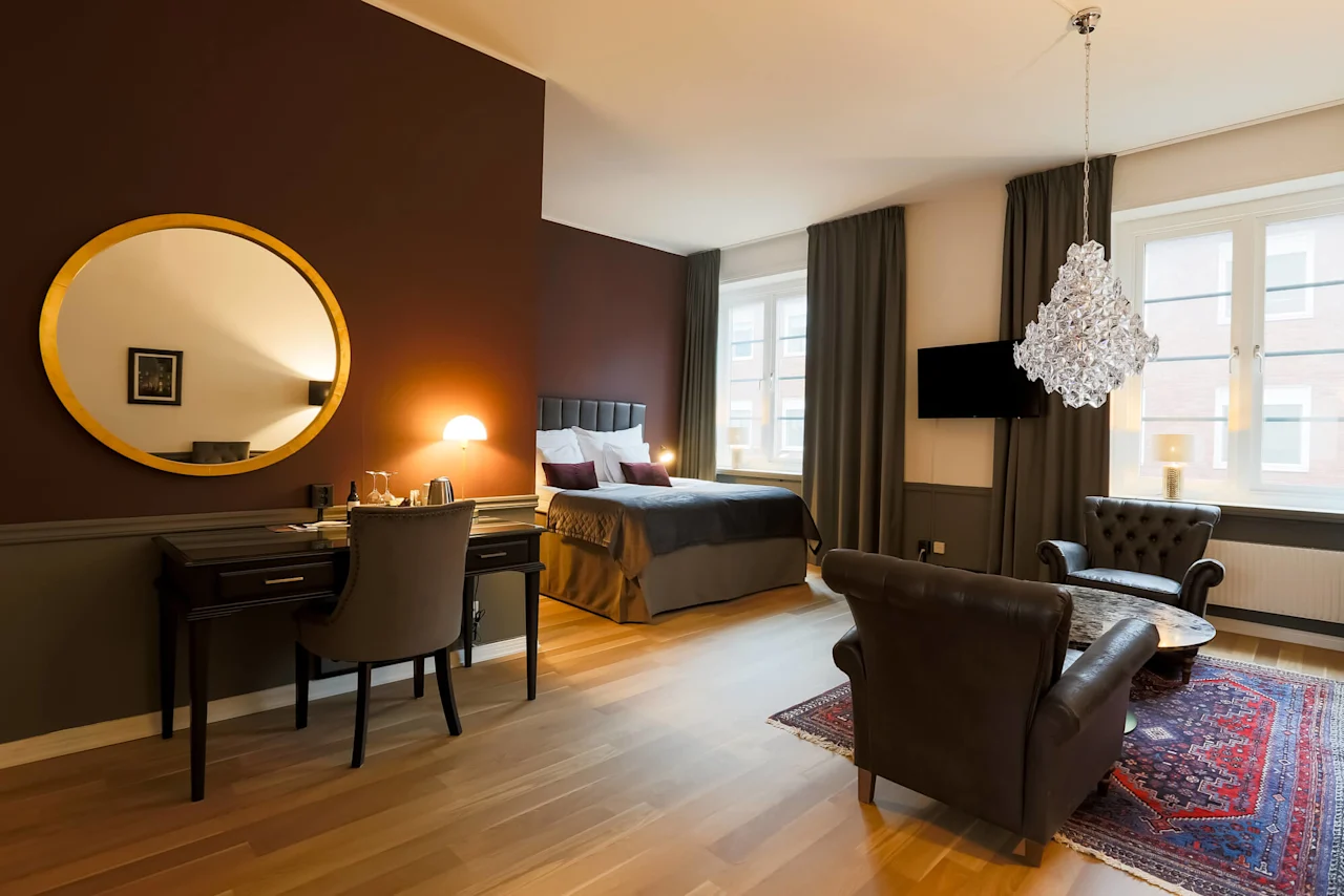 A well-appointed room with a large bed, desk, chairs, a chandelier, and a round mirror on a colored wall, suggesting a luxurious interior setting.