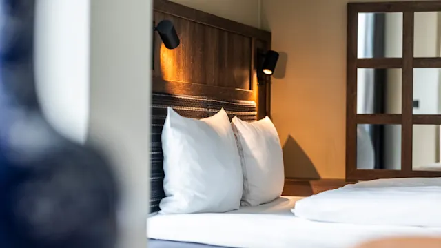 En pent oppredd seng med hvite puter er opplyst av varme nattbordslamper, mot en trehodegavl i et koselig, moderne rom.