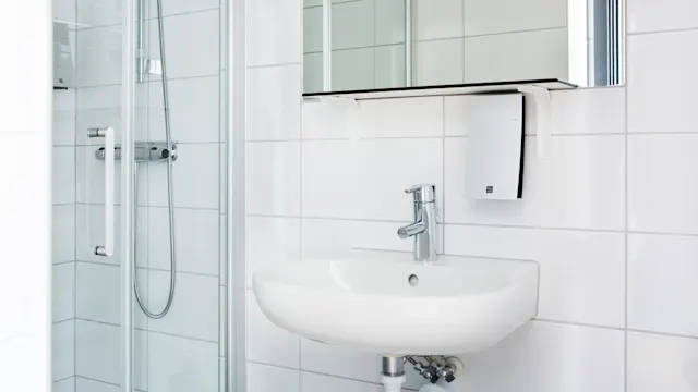 Handfat med kromblandare är installerat på en vit kakelvägg i badrummet. En glasdusch med silverhandtag och slang är bredvid.