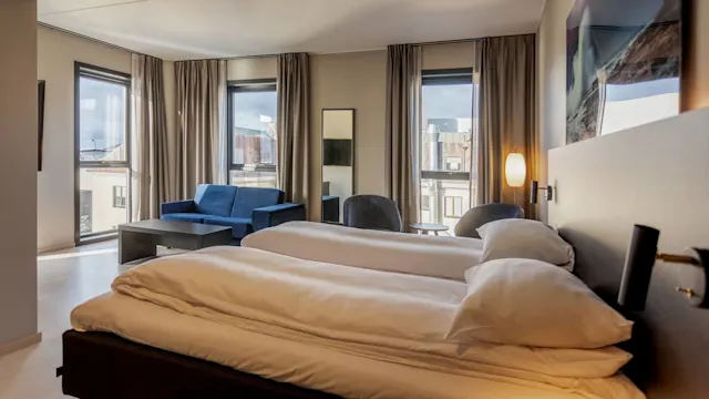 Et pent arrangert hotellrom med en dobbeltseng, hvitt sengetøy, en blå sofa, lenestol og vinduer med utsikt over bygninger. Naturlig lys lyser opp det moderne, romslige interiøret.