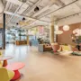 Lys, moderne lounge med fargerike stoler og bord arrangert avslappet. Overhengende pendellamper lyser opp trepanelvegger dekorert med planter. Neonskilt tilfører liv i en luftig, romslig setting.