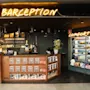 En bar med upplyst skylt "BARCEPTION" ovanför och en "COMFORT B" skylt till höger. Tidningsställ och ett kylskåp med snacks ingår i miljön.