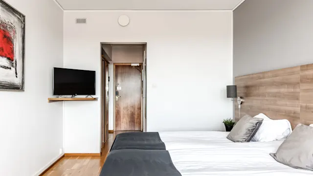 En säng med grå sängkläder är placerad i ett minimalistiskt rum. En väggmonterad TV och abstrakt konst dekorerar väggarna, med en trädörr i bakgrunden.
