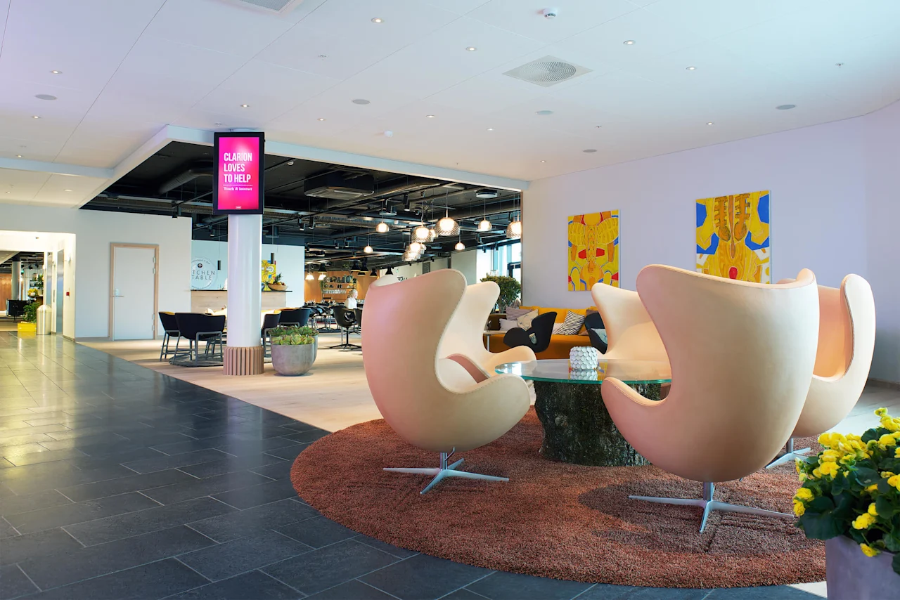 Fyra moderna stolar omger ett glasbord på en matta, i en stilren lobby med abstrakt konst. En skylt lyder: "CLARION LOVES TO HELP Travel & Business."