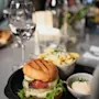 Burger med salat, ost, bacon og sylteagurker på en tallerken ved siden av pommes frites og saus. Et glass vann og en flaske i en svakt opplyst restaurant.