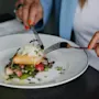 En tallerken med kokt fisk toppet med grønt serveres med grønnsaker. En person med røde negler skjærer den med bestikk ved siden av et glass hvitvin på et bord.