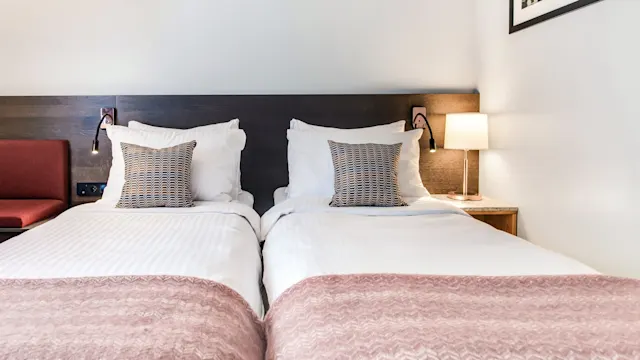To pent oppredde enkeltsenger med mønstrede puter står ved siden av hverandre. Hver seng har en leselampe over sengegavlen. Et innrammet bilde og en liten bordlampe pynter rommet.