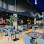 Moderne restaurantinteriør med blå stoler rundt marmorbord. Neonskilt lyser i bakgrunnen, mens potteplanter og omgivelseslys skaper en koselig atmosfære. Tekst inkluderer "barception" og "concession".