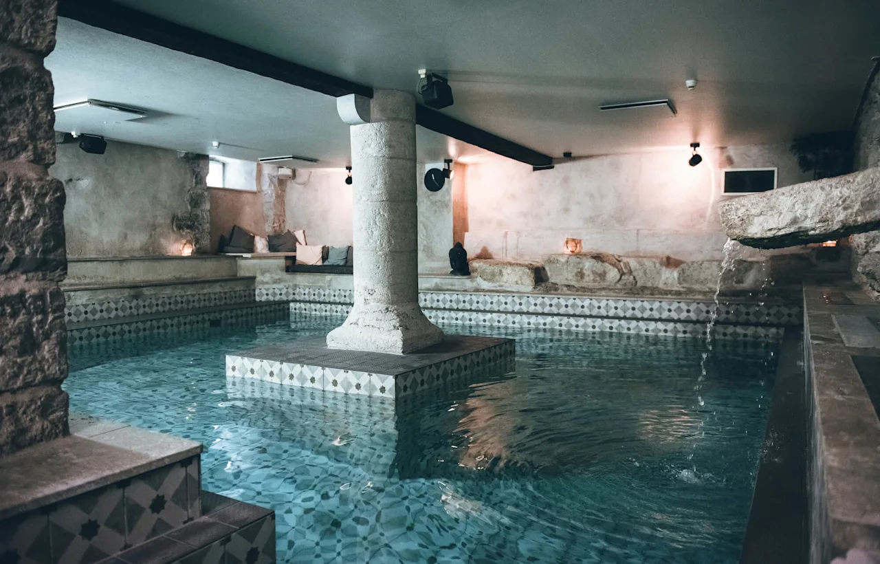 A stone column rises from a serene, tile-patterned indoor pool, surrounded by soft lighting and stone walls, with water gently cascading from a spout.