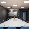 Hvitt konferansebord omgitt av grå stoler med papir og penner i et svakt opplyst rom med projektor, dekorativ vase og glassvegg.