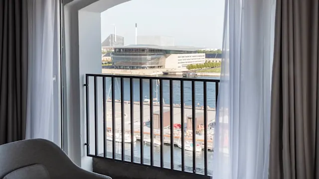 En balkongfönster med utsikt över en hamn ramar in en modern arkitektonisk byggnad med båtar vid en kaj under en klar himmel.