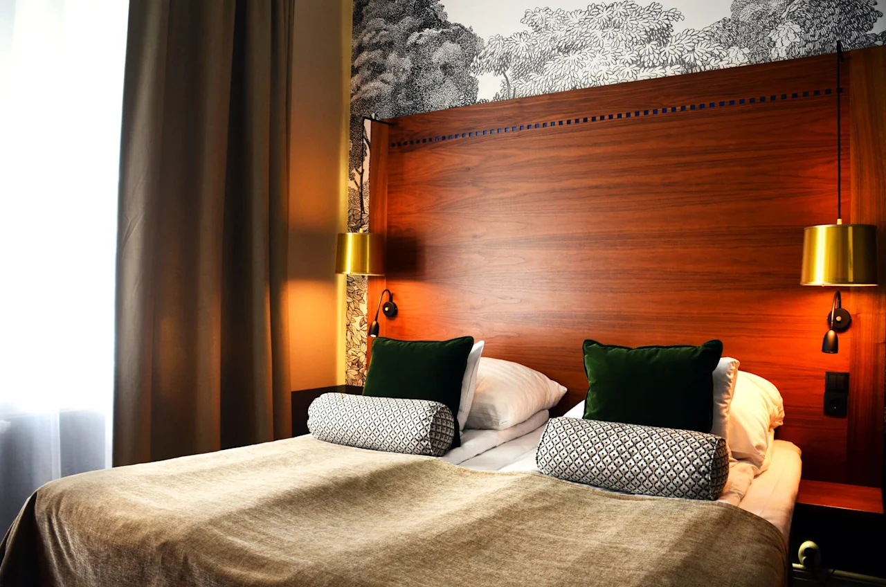 A cozy, neatly-made bed with pillows, flanked by brass wall lamps, against a wooden headboard and decorative wallpaper in a dimly lit room.
