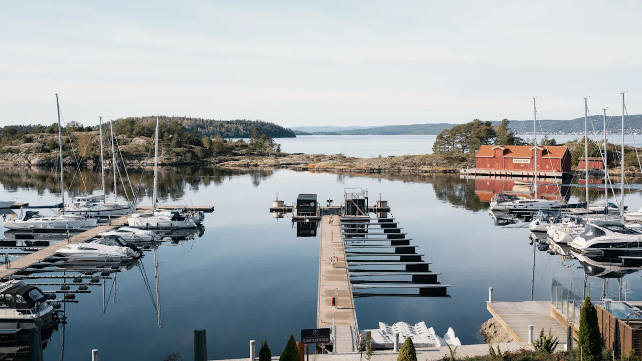 Både fortøjet ved en marina, omgivet af roligt vand og klippefyldt landskab. Et rødt bådehus med skilte: "Sørenga", "Kafé & Bar", "Gjesteplass", og "GjesteBrygge."