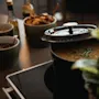 En gryte med suppe simmer på en komfyr, delvis lokket, pyntet med urter. I nærheten står skåler med frø og stekte grønnsaker, og en plante dekorerer scenen.