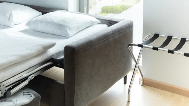 En grå bäddsoffa står bredvid ett stort fönster som vetter mot en gata med träd och byggnader utanför. En bagageställning står nära på trägolvet.