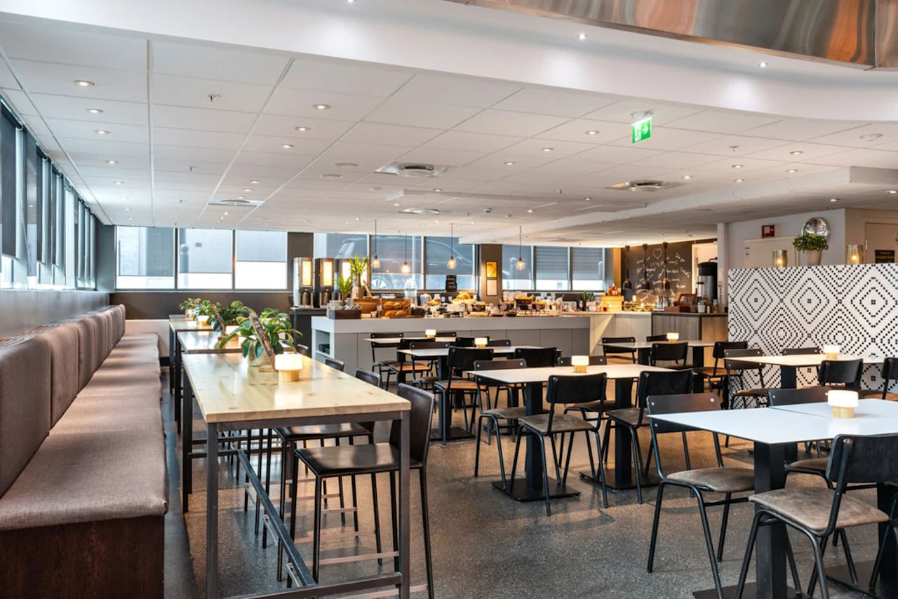 En rymlig restauranginteriör har ordnade bord och stolar. Ljus står på borden och skapar stämning. Stora fönster släpper in naturligt ljus, och bufféområdet är synligt i bakgrunden.