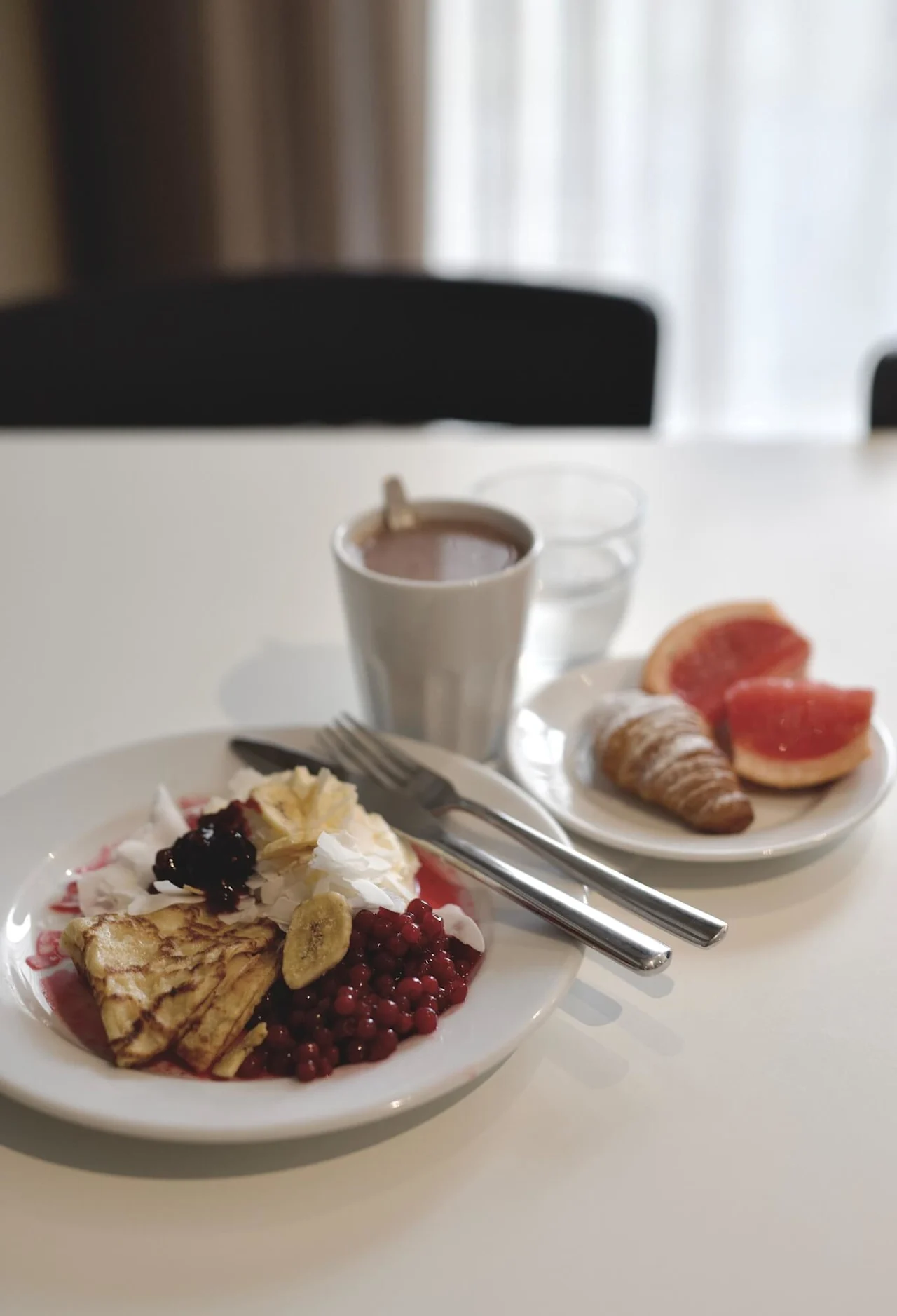 En frokostdekning med pannekaker, bær og fruktskiver på en tallerken, en kopp varm drikke og bestikk, satt på et hvitt bord i et mykt opplyst rom.