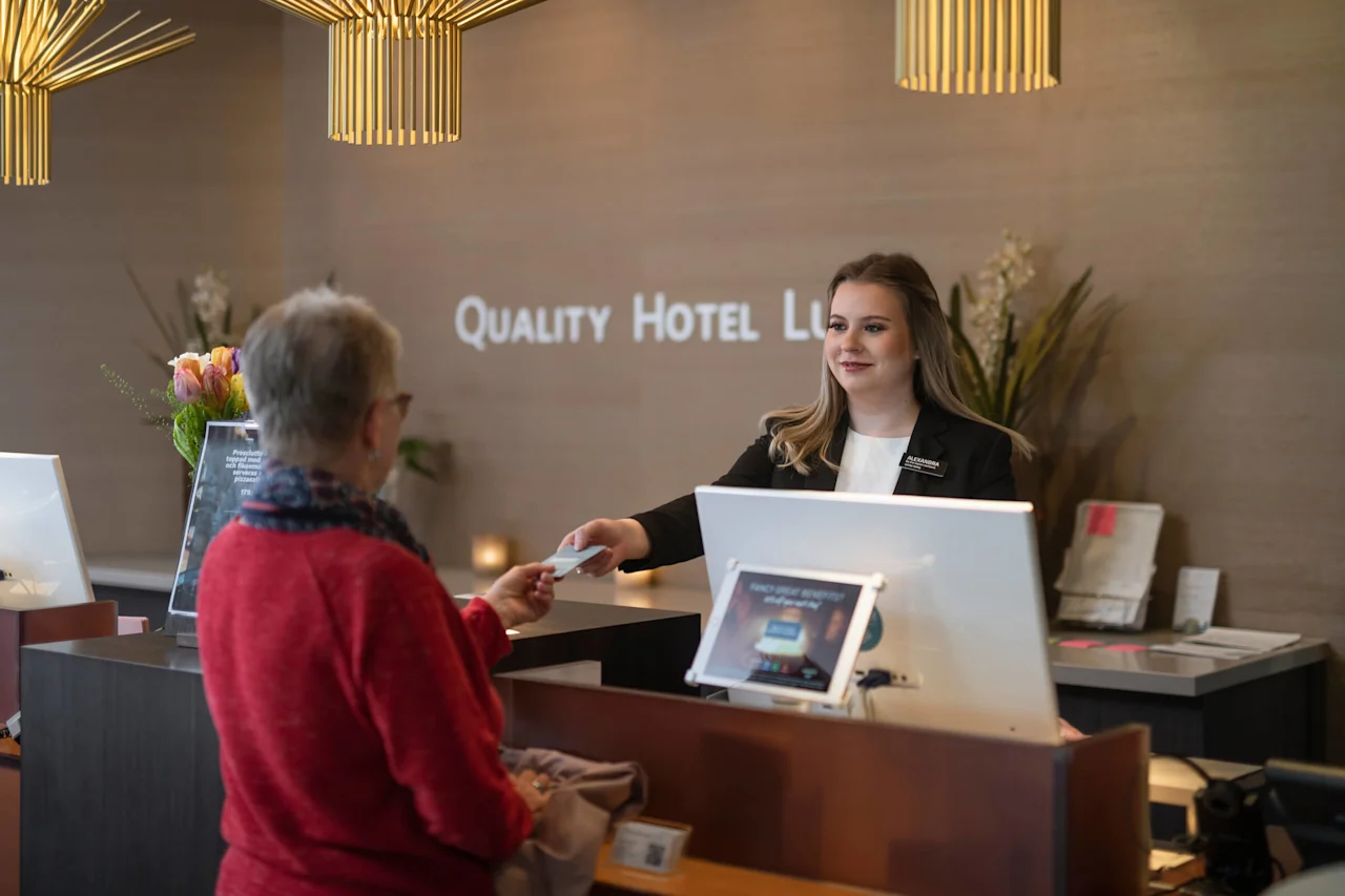 Receptionist handing a card to a guest at a hotel check-in desk. Elegant lighting above, flowers nearby. Sign reads "QUALITY HOTEL LLE.” Reception feels modern and welcoming.