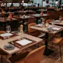 Bord dekket for middag, med vinglass, tallerkener og brettede servietter, skaper en koselig atmosfære i en varmt belyst restaurant med tregulv og pent arrangerte stoler.