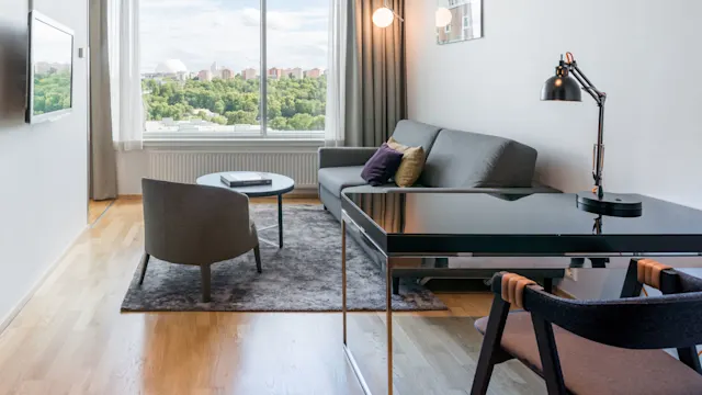 Ett vardagsrum med en grå soffa med färgglada kuddar och ett runt bord under ett stort fönster med utsikt över stadsvyn. En svart skrivbordslampa står på ett elegant skrivbord.