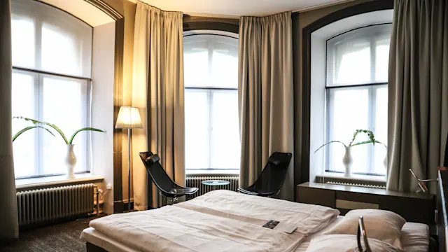En seng med hvidt sengetøj står i et lyst rum med store vinduer, flankeret af to sorte stole, en lampe og potteplanter på vindueskarmene.