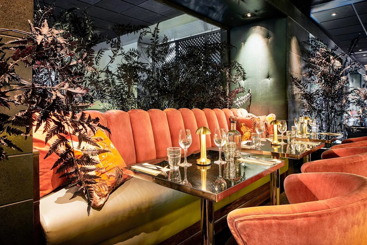 Polstrede laxrosa sitteplasser med livlige puter omgir et stilfullt spisebord med elegant glass og en gullampe, innrammet av frodig, mørk vegetasjon i en svakt opplyst restaurant.