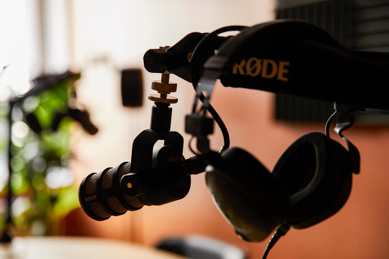 Mikrofon på en justerbar arm är placerad över ett bord. Hörlurar ligger i närheten. Oskarpa växter och akustikpaneler i bakgrunden. Text: "RØDE" på armen, "RODE" på mikrofonen.