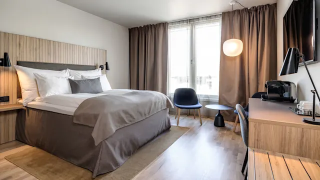En seng med hvitt og grått sengetøy står ved et vindu med brune gardiner. En stol og et bord står i nærheten, og et skrivebord med kaffemaskin fullfører rommet.