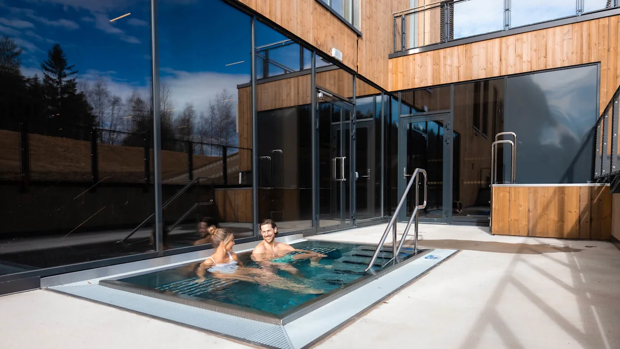 To personer slapper av i et utendørs boblebad ved siden av en moderne trebygning under en klar blå himmel på Lily Country Club.