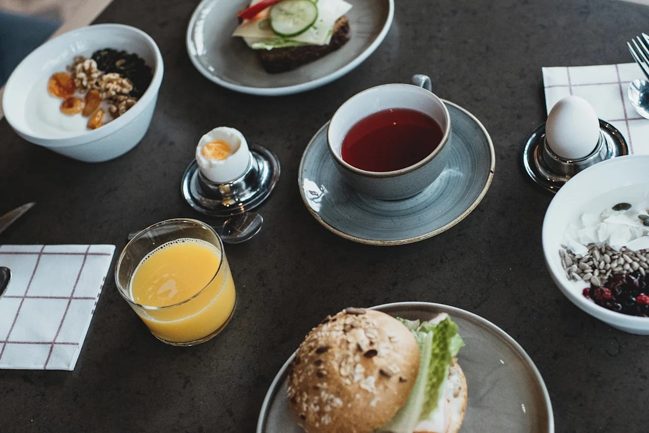 En frokostoppsett med en sandwich, yoghurtskåler med nøtter og bær, et bløtkokt egg, en kopp te og et glass appelsinjuice på et mørkt bord.