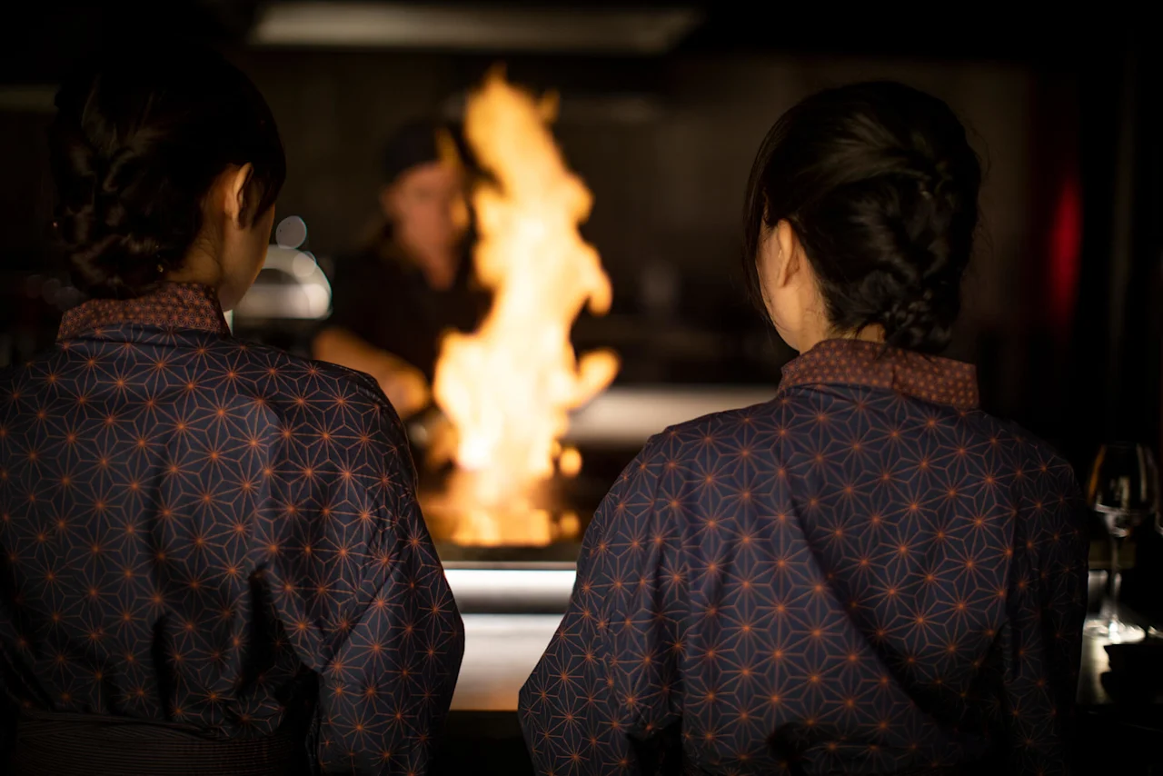 To personer i mønstrede uniformer ser på en kokk som lager mat med flammer, på et restaurantkjøkken.