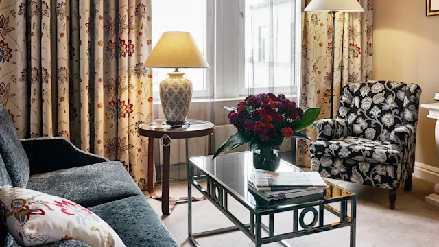 En stue med en blomstrete lenestol og sofa, et lampet bord, et salongbord med blomster og magasiner, alt satt mot blomstrete gardiner og et vindu.