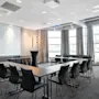 Konferencelokale med U-formet bordopstilling, tomme stole, projektor og naturligt lys fra store vinduer.