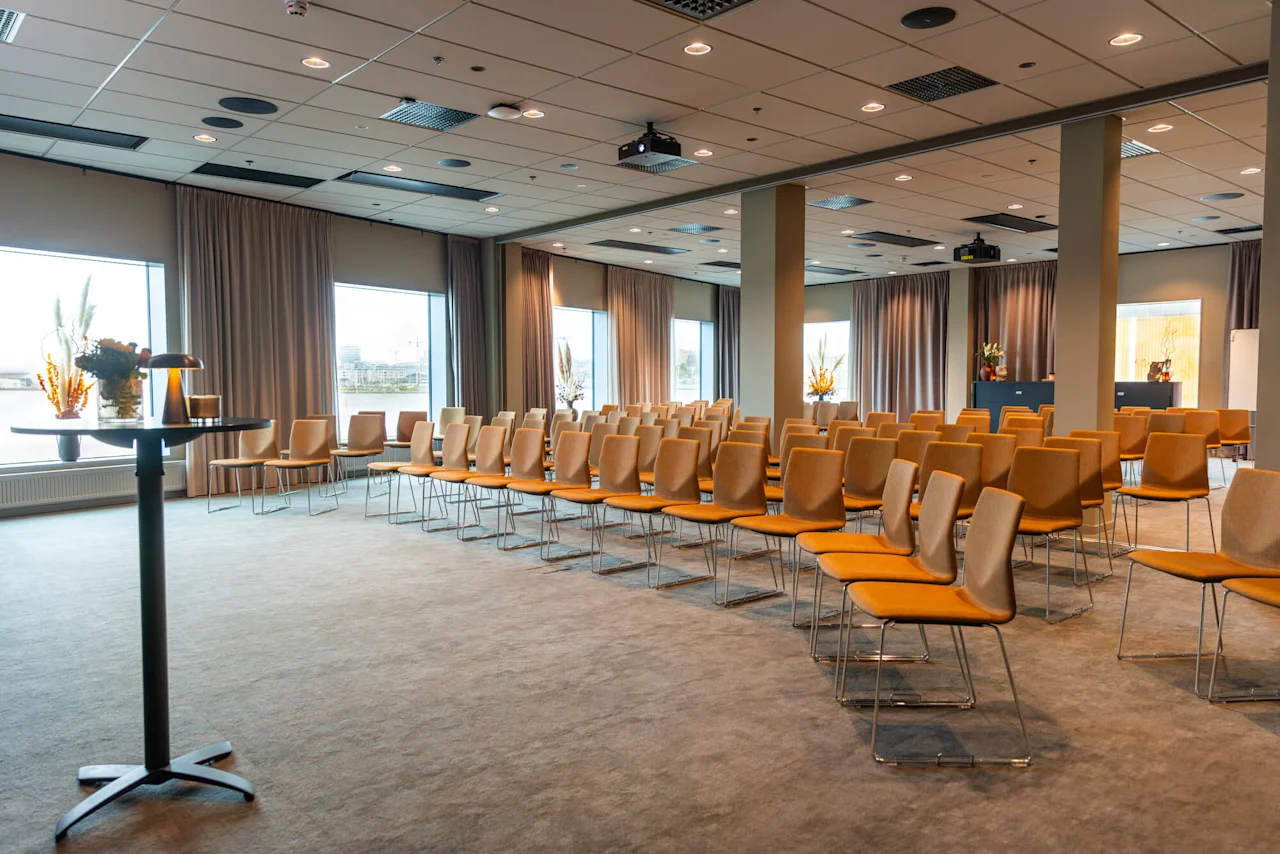 Rækker af orange polstrede stole er arrangeret i et konferencelokale. Store vinduer med gardiner og potteplanter skaber en åben, luftig atmosfære.
