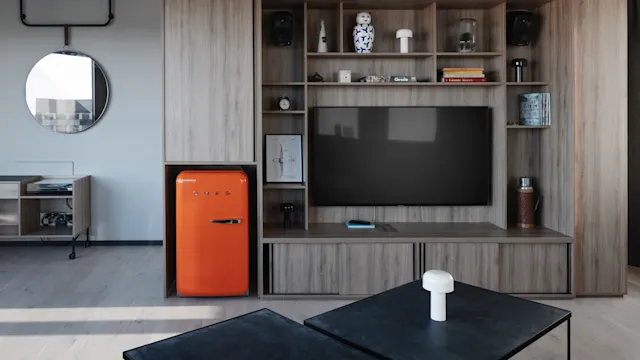 A wooden entertainment unit with shelves holds decorative items and speakers. An orange SMEG fridge is on the left. A large TV is centered. A coffee table sits in front.