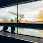To personer slapper av i et innendørs basseng, og ser ut gjennom et stort vindu som viser et montert jagerfly og høsttrær under en skyet himmel.