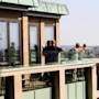A group of people relaxes on a glass-paneled balcony of a modern building, overlooking a cityscape with a river and distant hills under a clear sky.