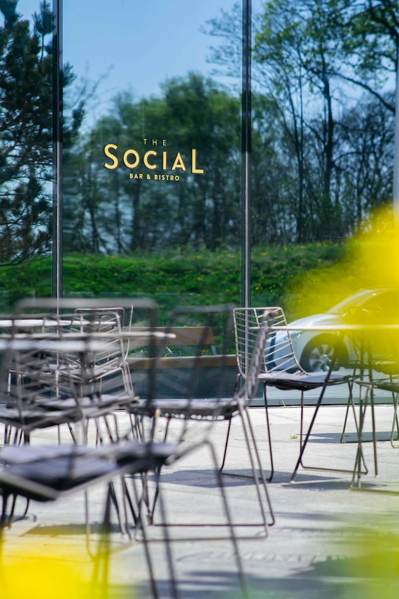 Empty metal chairs and tables stand on a patio. Reflective glass shows trees. Yellow abstract blur is in the foreground. Text on glass: "THE SOCIAL BAR & BISTRO". 