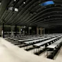 Lange borde med stoler er arrangert i rader mot et podium i et romslig, mørkt takoverdekket konferanserom. Veggene er dekket med vinduer og gardiner under omgivelsesbelysning.