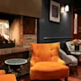 En hyggelig lounge med en brændende pejs indbygget i en murstensvæg, omgivet af orange og mønstrede stole, små borde, trægulv, kunstværker og dæmpet belysning, der skaber en varm atmosfære.