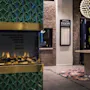 Elektrisk peis med realistiske flammer på en grønn, mønstret vegg. Murvegg og et skilt med teksten "EGON RESTAURANT" i bakgrunnen. Rundt bord med blomster under en dekorativ lysekrone.