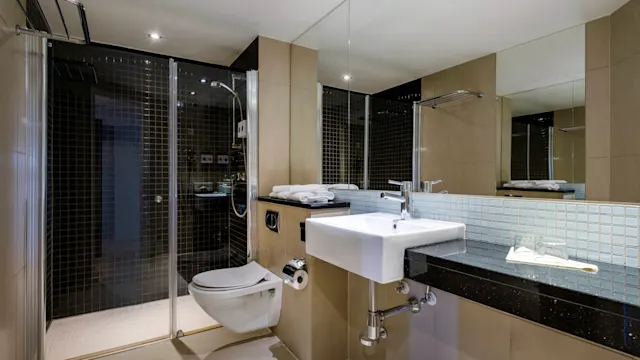 Et moderne bad har en veggmontert vask og toalett med en innglasset dusj ved siden av. Konteksten viser glatte, reflekterende overflater, og nøytrale fliser forsterker den moderne designen.