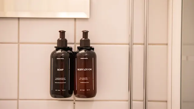 To vægmonterede flasker mærket "SOAP" og "BODY LOTION" med pumper, i et flisebelagt badeværelse. Teksten på hver lyder, ”MADE NOTES” og ”Hand Made”. Flaskerne er brune.