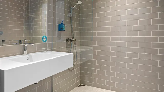 Elegant hvit vask med en metallkran i et flislagt bad. I nærheten, en dusj med glassvegg. Veggmontert pumpeflaske merket: "DOVE MEN+CARE".