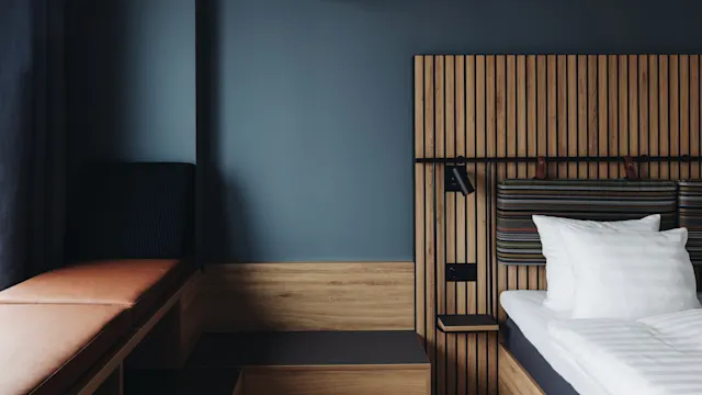 A modern bedroom features a wooden bed with white pillows and a slatted headboard, next to a leather bench under a spherical ceiling light, set against dark walls.